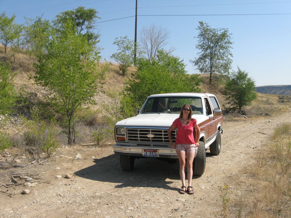 Woman in pink shirt and shorts stands by a 1980 Ford Bronco on a dirt road. License plate reads 'IA SK476'