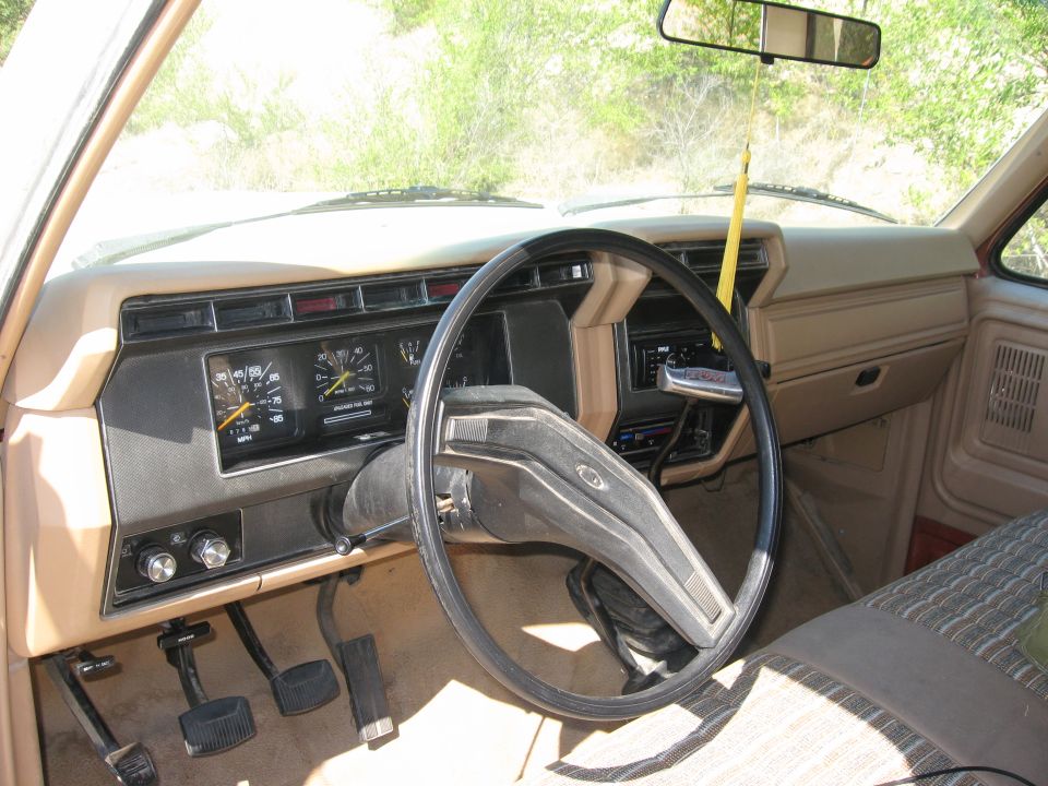 Interior view of a 1980s Ford truck dashboard with steering wheel, gauges, and pedals visible. The dashboard is tan