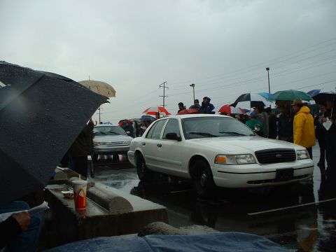 1999 Ford Crown Victoria - A white sedan is parked at an outdoor auction on a rainy day with many people holding umbrellas.