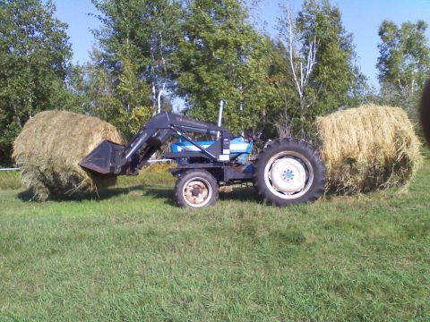 1987 Ford F-250 - A blue tractor with a front loader is positioned between two large round hay bales in a grassy field.
