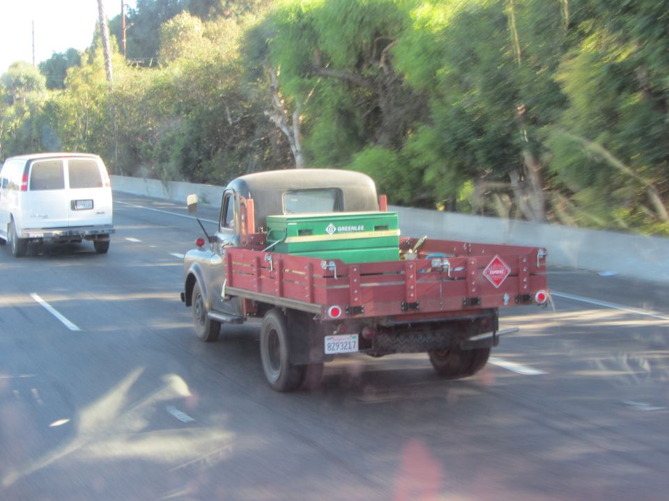 Old flatbed truck with red wooden sides and a green toolbox in the bed, driving on a highway next to a white van