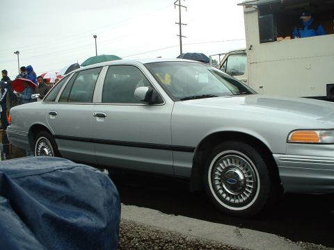 1999 Ford Crown Victoria - A silver 1999 Ford Crown Victoria is parked on a wet surface with people holding umbrellas in the background.