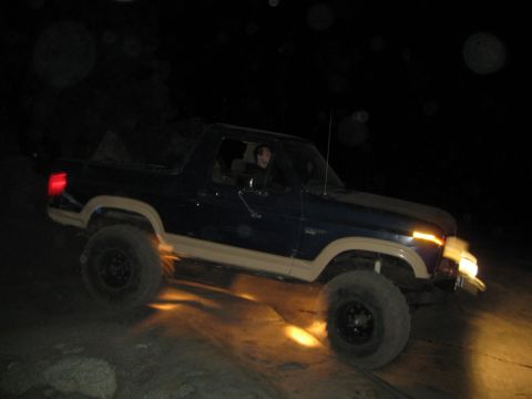 1986 Ford Bronco - A dark blue Ford Bronco with tan trim is driven off-road at night, its headlights illuminating the dusty path.