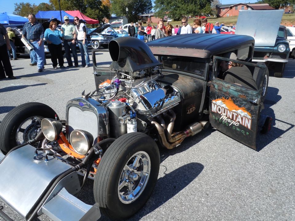 Rat rod with exposed engine, scoop, and "Mountain Magic Speed Shop" door art. Other cars and people in background