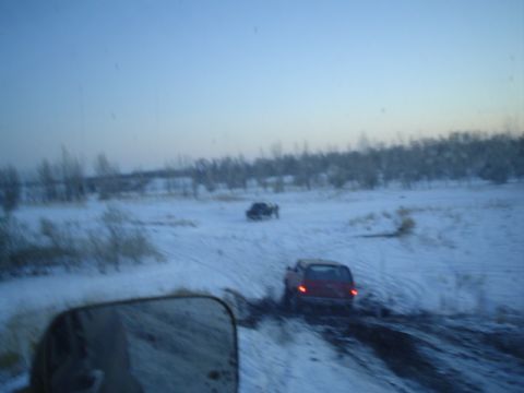 1981 Ford Bronco - Dusk shot taken over a side mirror of a red SUV with brake lights lit, working through a muddy rut in a snow-covered field, with a second d…