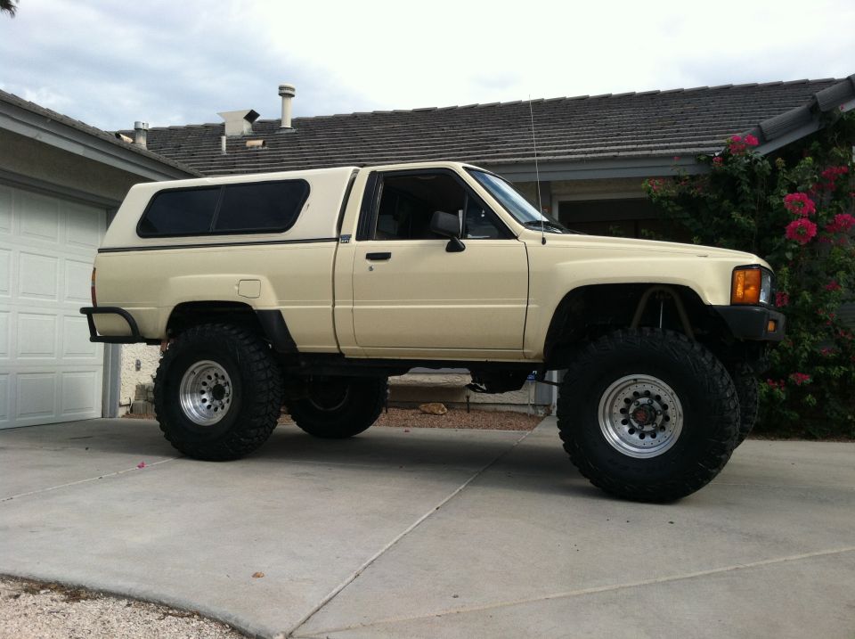 Cream-colored Toyota pickup truck with a camper shell, lifted with large off-road tires, parked on a driveway