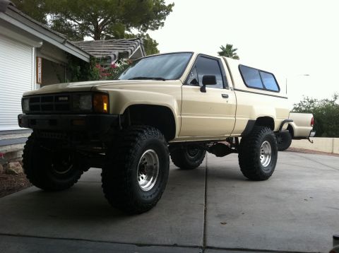1985 Toyota Pickup/SR5 - This lifted beige pickup truck with a camper shell is shown from a 3/4 front angle, parked in a driveway.