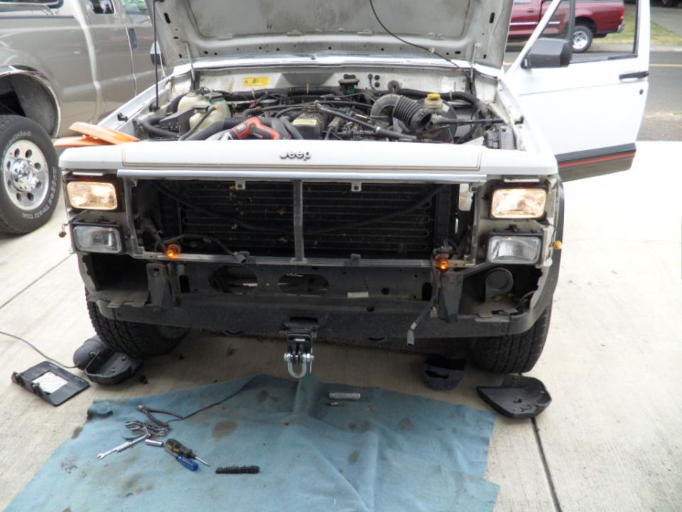 1994 Jeep Cherokee front end with hood open, showing engine and grille. Bumper is partially removed. Another vehicle is visible to the left