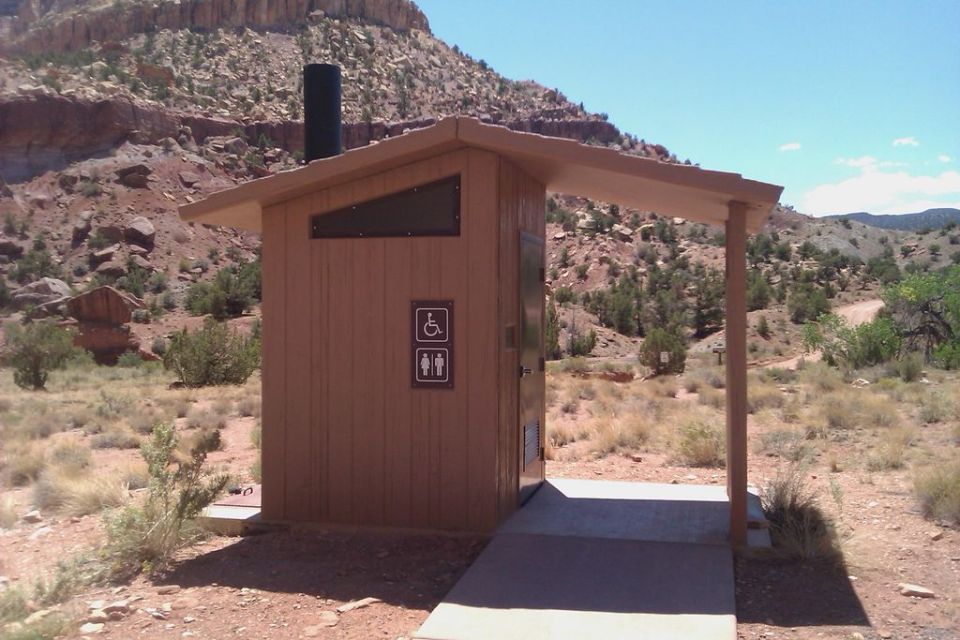 Wooden public restroom building with a black chimney and accessibility symbols, set in a dry, rocky landscape