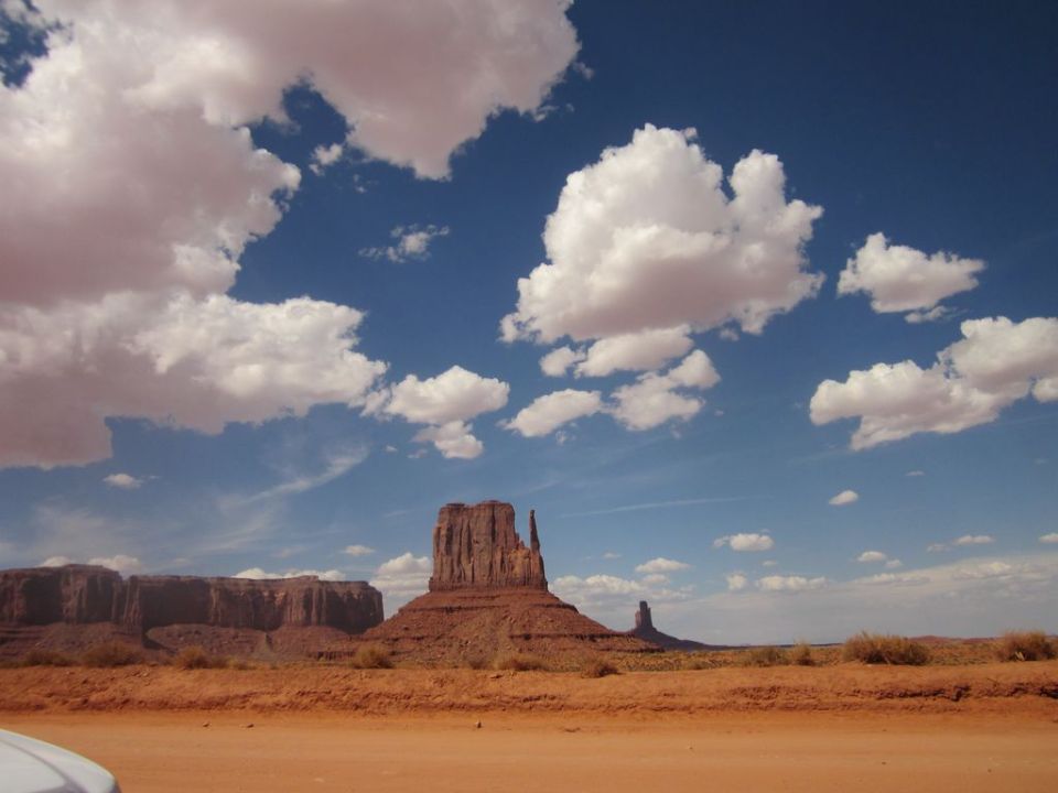 Monument Valley landscape with buttes under a cloudy blue sky. Part of a white vehicle in the foreground