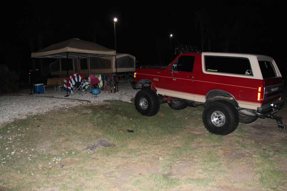 1990 Ford Bronco parked at a campsite at night, lifted suspension, red exterior with tan trim