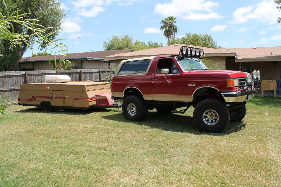 1990 Ford Bronco with lifted suspension and off-road lights, towing a tan pop-up camper
