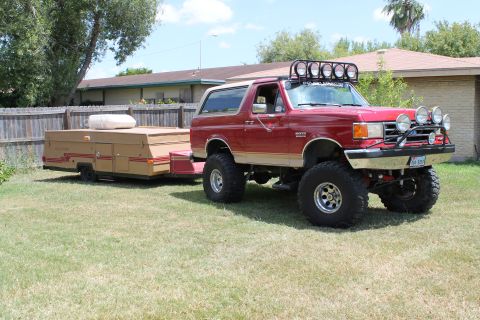 1990 Ford Bronco - A lifted red Ford Bronco tows a pop-up camper in a grassy yard.
