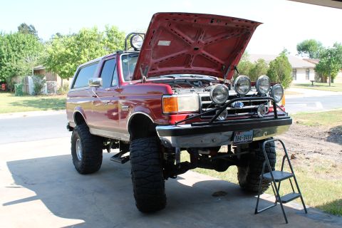 1990 Ford Bronco - This lifted red Ford Bronco has its hood open, revealing the engine bay and extra lights mounted on the front grille guard.