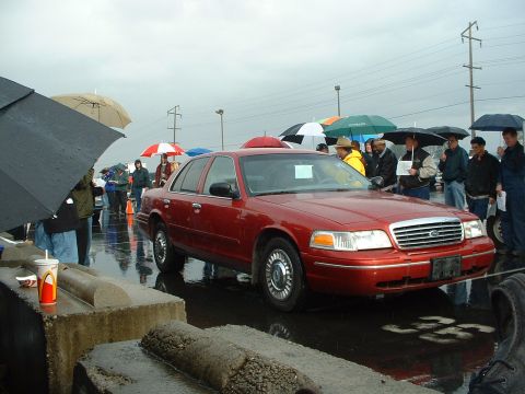 1999 Ford Crown Victoria - A red 1999 Ford Crown Victoria is displayed at an outdoor auction under rainy skies with several people holding umbrellas.