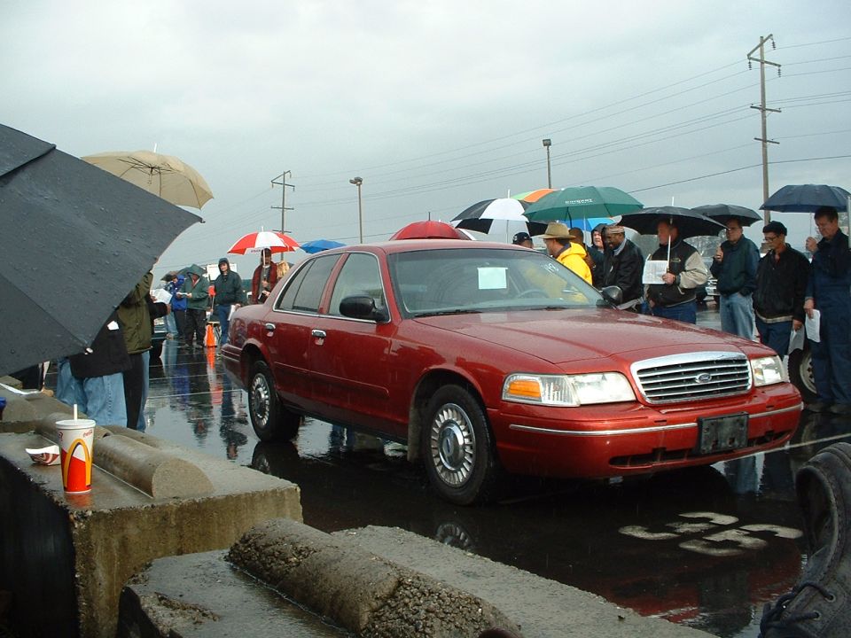 Red Ford Crown Victoria sedan at a rainy outdoor auto auction with bidders under umbrellas