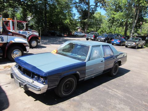 1991 Ford LTD Crown Victoria - A two-tone blue sedan, possibly a 1991 Ford Crown Victoria, sits in a parking lot with other vehicles and tow trucks.