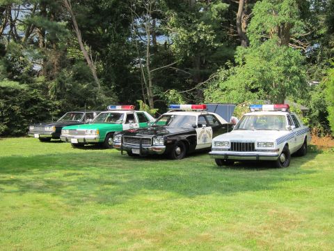 1991 Ford LTD Crown Victoria - Four vintage American police cars lined up on a shaded lawn, including green-and-white, black-and-white and dark-colored full-size sedans.