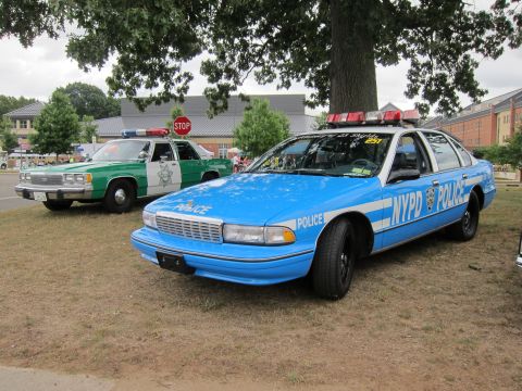 1991 Ford LTD Crown Victoria - light-blue Chevrolet Caprice marked NYPD POLICE displayed on grass beside a green-and-white police sedan near a STOP sign.