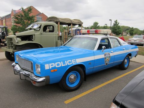 1991 Ford LTD Crown Victoria - light blue Plymouth police sedan with roof light bar parks beside an olive military truck at an outdoor car show.