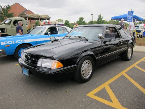 1991 Ford LTD Crown Victoria - black Ford Mustang LX notchback coupe with 5.0 badge parks beside a light blue police car at an outdoor show.