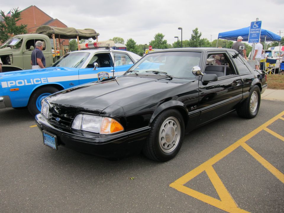 Black Ford Mustang LX coupe parked beside a blue police car at a car show