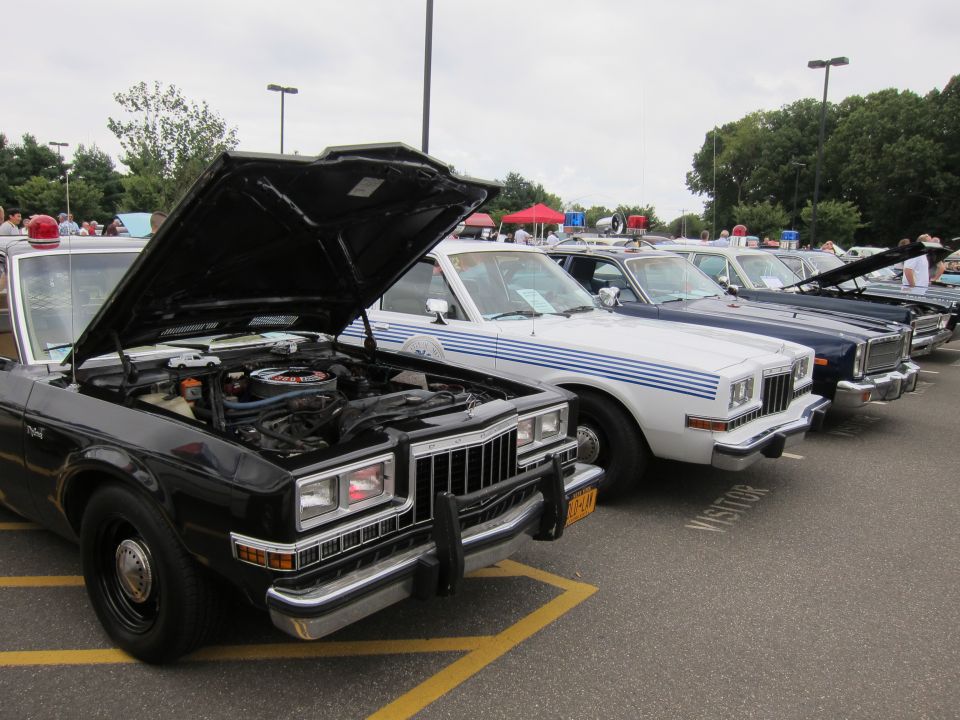 Row of classic police sedans, one black with hood open, in a parking lot show