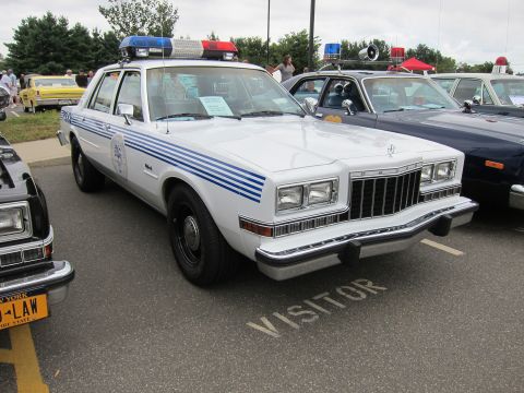 1991 Ford LTD Crown Victoria - white police sedan with blue side stripes, chrome grille and roof light bar parks over a VISITOR-marked space at a show.