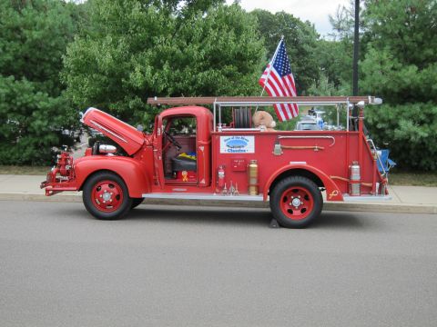 1991 Ford LTD Crown Victoria - red vintage fire engine pumper with raised hood, ladder rack and a U.S. flag parks along a curb beside greenery.