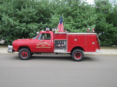 1991 Ford LTD Crown Victoria - red Dodge 4x4 fire pumper lettered Central Fire Co. with a U.S. flag and ladder rack parks along a street curb.