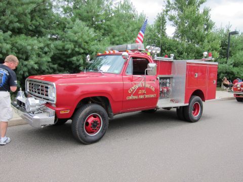 1991 Ford LTD Crown Victoria - man stands beside a red Dodge 4x4 fire pumper lettered Central Fire Co. with U.S. flag and ladder rack at the curb.