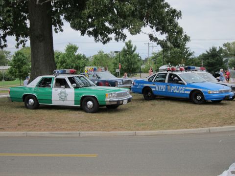 1991 Ford LTD Crown Victoria - Two vintage police cars: a green-and-white sheriff sedan beside a blue NYPD cruiser, parked on grass at a show.