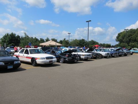 1991 Ford LTD Crown Victoria - long row of vintage police cars parked in a paved lot under blue sky, led by a white-and-red sheriff sedan.