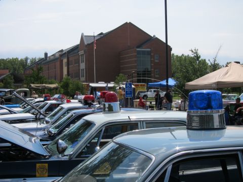 1991 Ford LTD Crown Victoria - lineup of vintage police cars with red and blue roof beacons parked at a show in front of a large brick building.