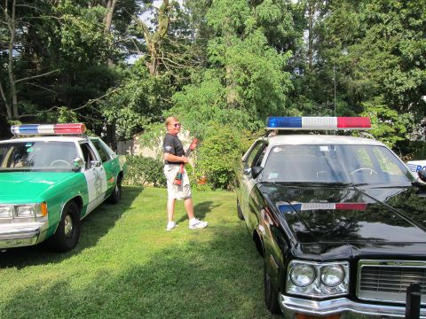 1991 Ford LTD Crown Victoria - man stands between a green-and-white sheriff car and a black police cruiser parked on a grassy lawn near trees.