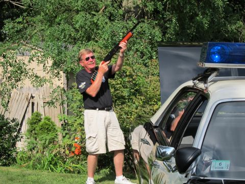 1991 Ford LTD Crown Victoria - man in a dark shirt and shorts raises a shotgun skyward while standing next to a police cruiser with a blue beacon.