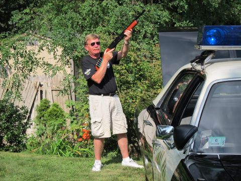 1991 Ford LTD Crown Victoria - man in sunglasses points a shotgun upward beside a police cruiser with a blue roof beacon, a wooden fence behind him.