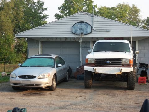 1989 Ford Bronco - A white 1989 Ford Bronco is parked in front of a garage, next to a silver sedan.