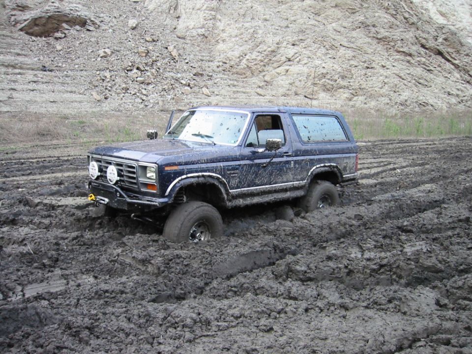 Muddy dark blue Ford Bronco SUV stuck in deep mud, front three-quarter view with brush guard and winch.