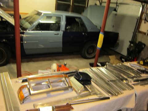 1991 Ford LTD Crown Victoria - A sedan, possibly undergoing restoration, sits in a garage with its hood and trunk open. Various trim pieces are laid out on a table in the…