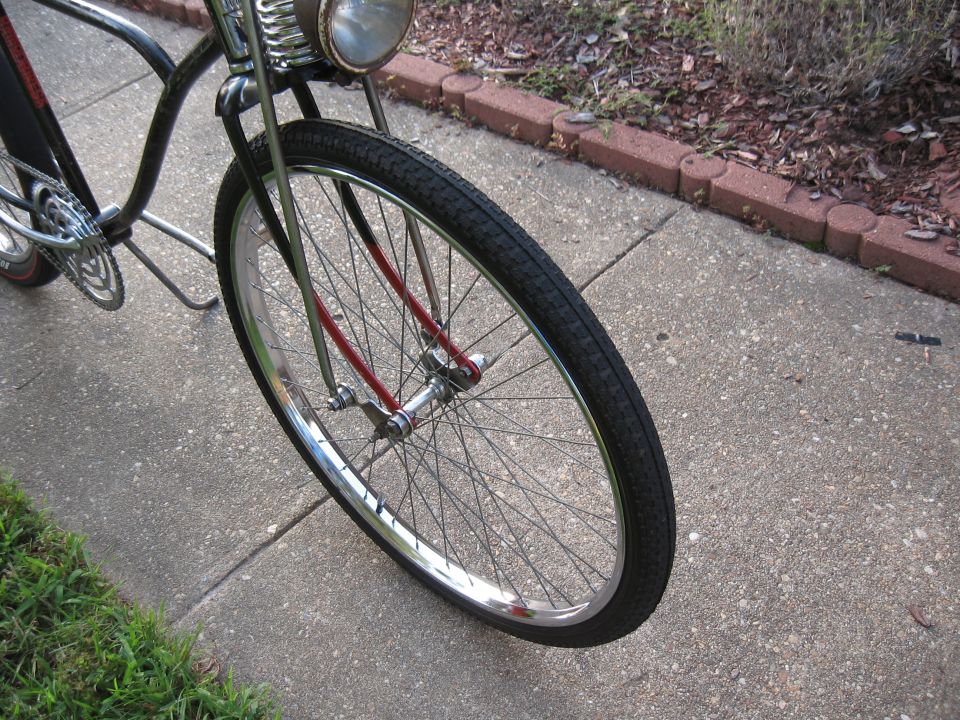 Close-up of a bicycle's front wheel and part of the frame. Chrome handlebars and a headlight are visible
