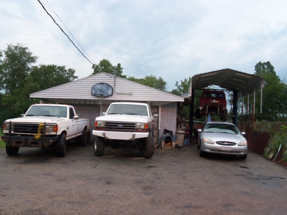 Two white Ford trucks and a silver sedan parked outside a house; a red truck is on a lift
