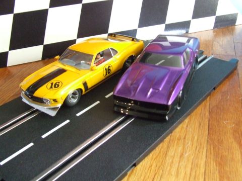 1973 Ford Mustang - Two slot cars, one yellow and one purple, are positioned on a black race track with white lane markers.