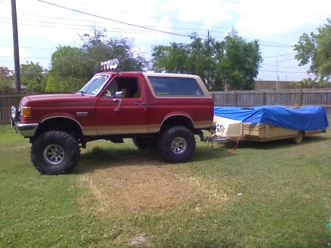 1990 Ford Bronco - A lifted red 1990 Ford Bronco is parked on a grassy lawn, hitched to a pop-up camper covered with a blue tarp.