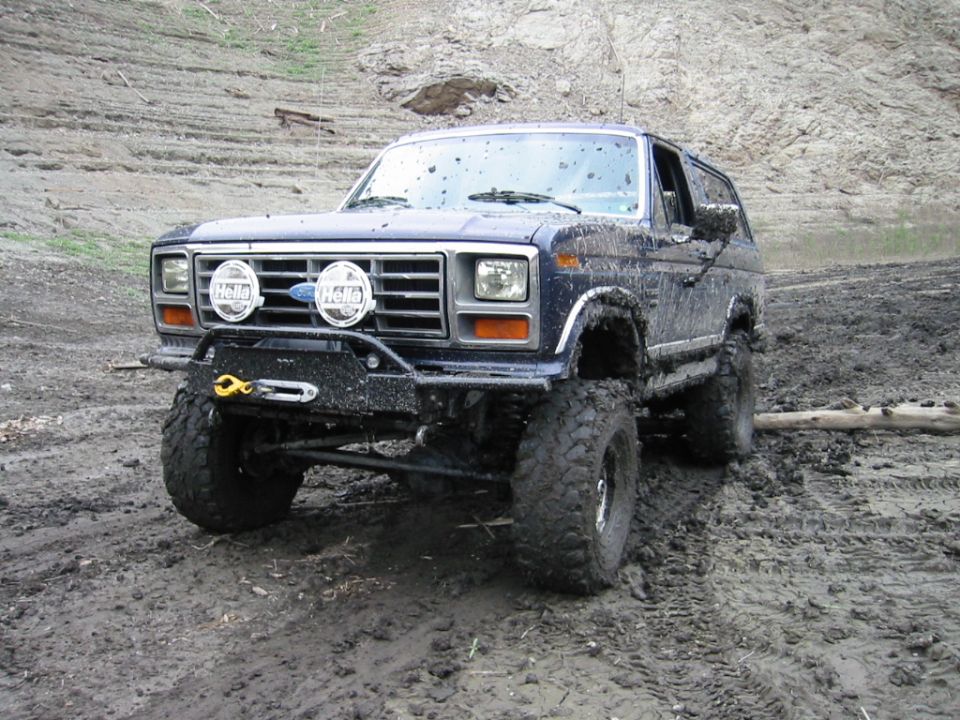 Lifted dark Ford Bronco on mud tires with KC lights and brush guard parked in a muddy off-road pit