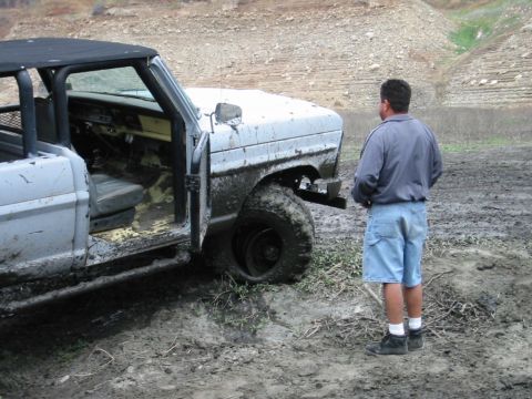 1985 Ford Bronco - A mud-covered 1985 Ford Bronco sits stuck in a muddy area, its driver's side door open, with a man observing the scene.