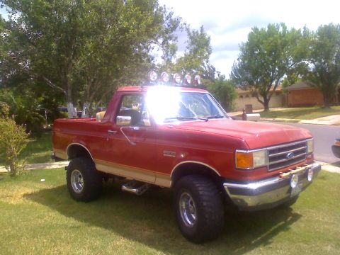 1990 Ford Bronco - This red Ford Bronco features off-road tires and a light bar, parked on a grassy lawn.