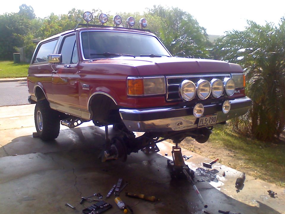 1990 Ford Bronco with front suspension removed, sitting on jack stands. Four driving lights are mounted above the bumper