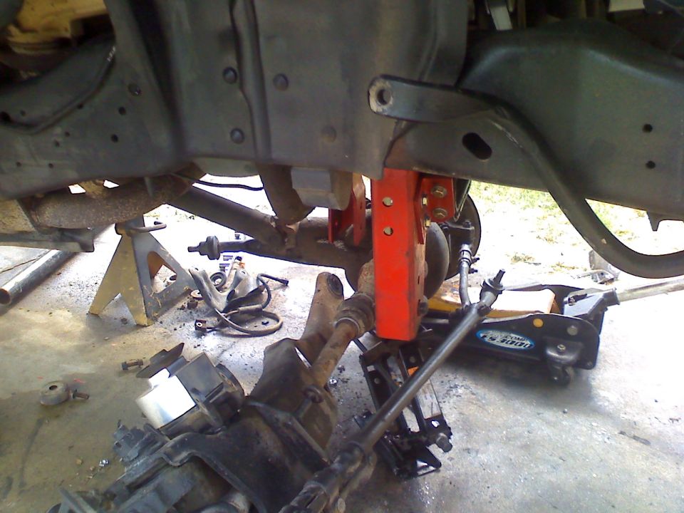 Undercarriage of a vehicle on jack stands, showing suspension components and a hydraulic jack