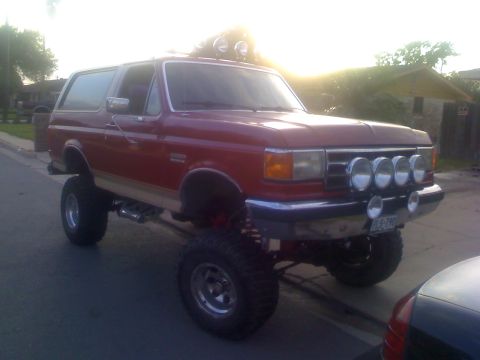 1990 Ford Bronco - A lifted red 1990 Ford Bronco is parked on the street with a bright sun in the background.